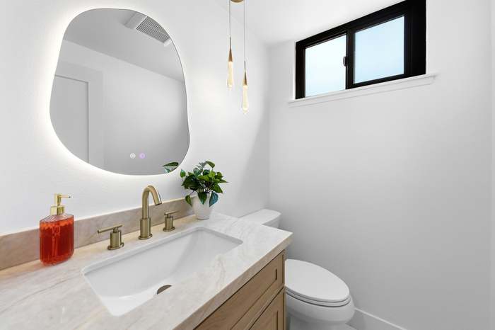 Powder room vanity with backlit organic-shaped mirror, brushed gold faucet, and pendant lighting