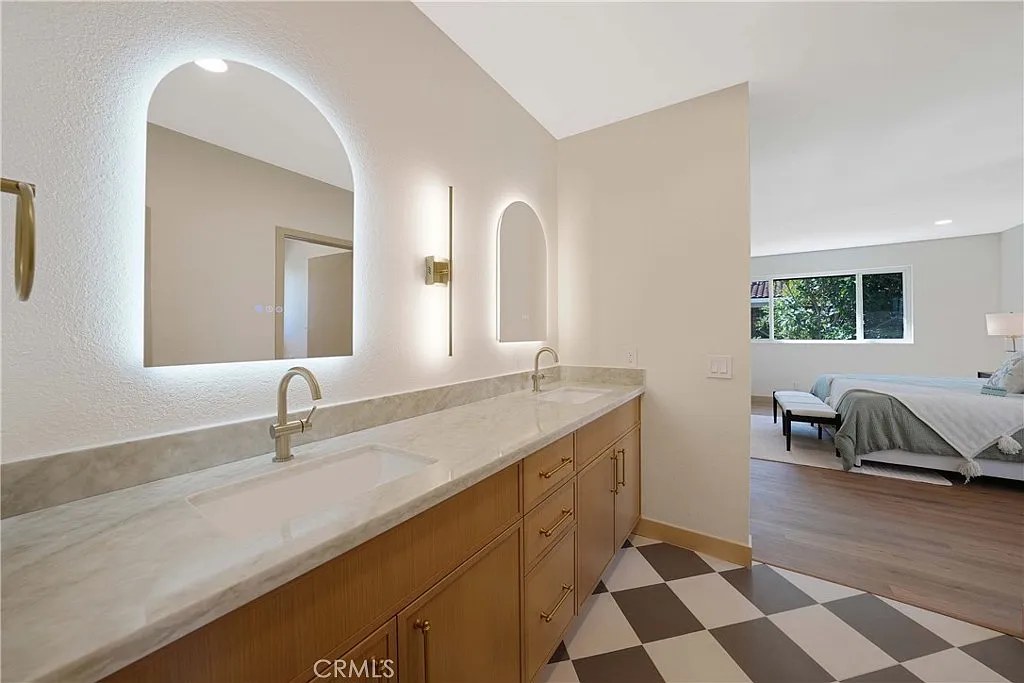Long double vanity with two backlit arched mirrors, marble countertop, and view into an adjoining bedroom