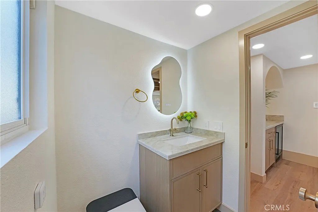 Powder room vanity with backlit organic-shaped mirror and brushed gold faucet, open to an adjoining bar area