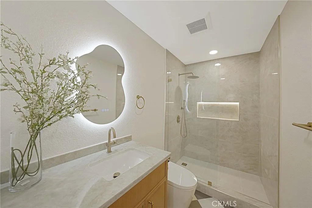 Bathroom vanity with backlit organic-shaped mirror, marble countertop, and view into a walk-in glass shower