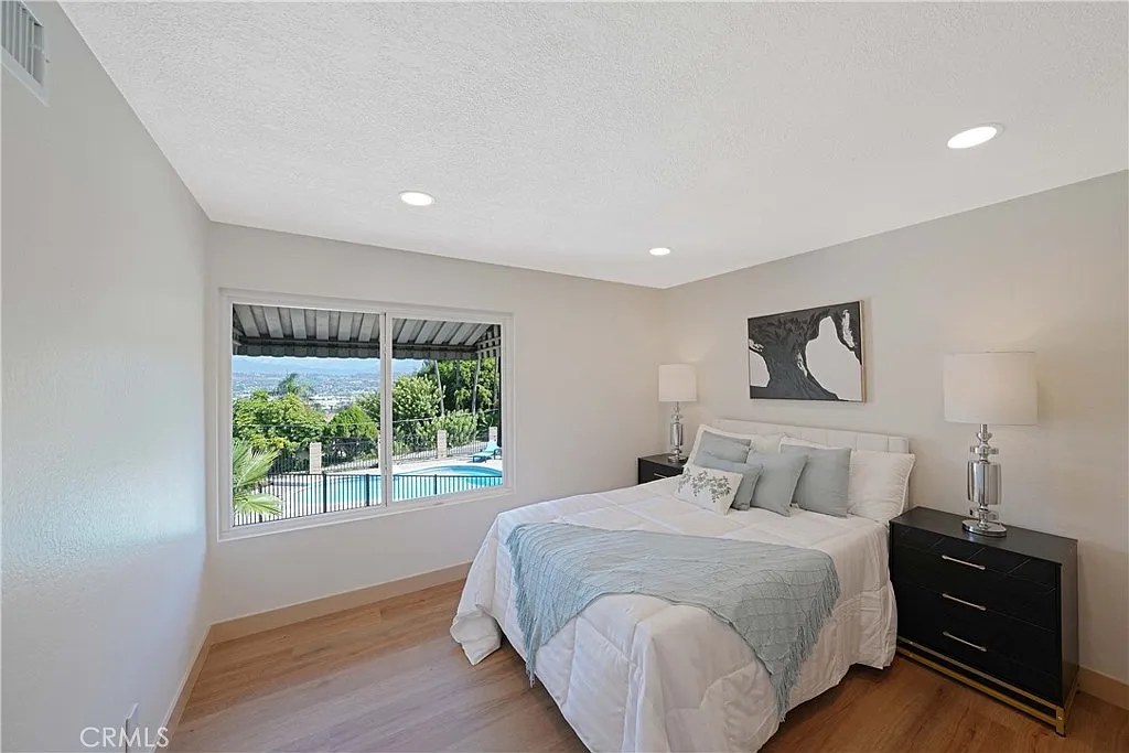 Bedroom with picture window overlooking a pool and city view, staged with light bedding