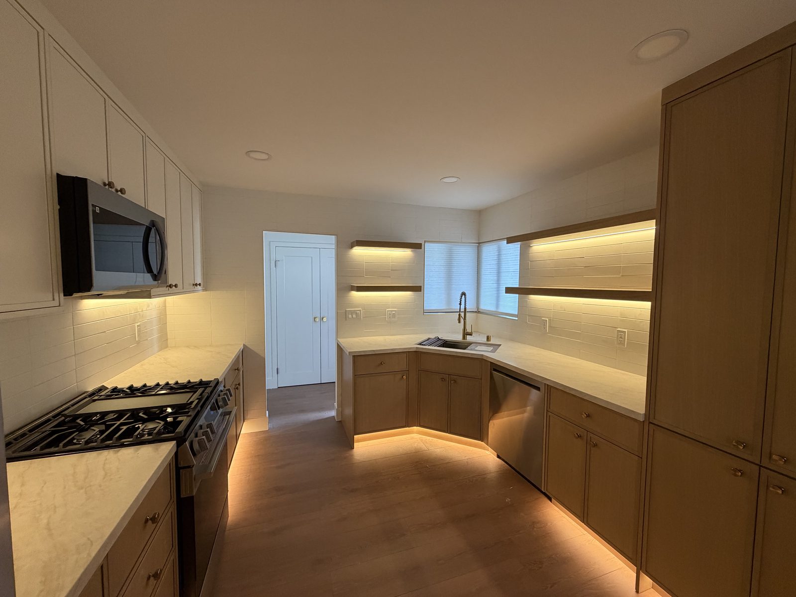 Two-tone white and wood kitchen with corner sink, gold faucet, and floating shelves with under-cabinet lighting