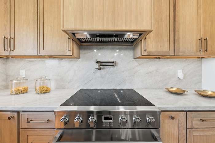 Close-up of a stainless range with pot filler, marble backsplash, and light wood cabinetry
